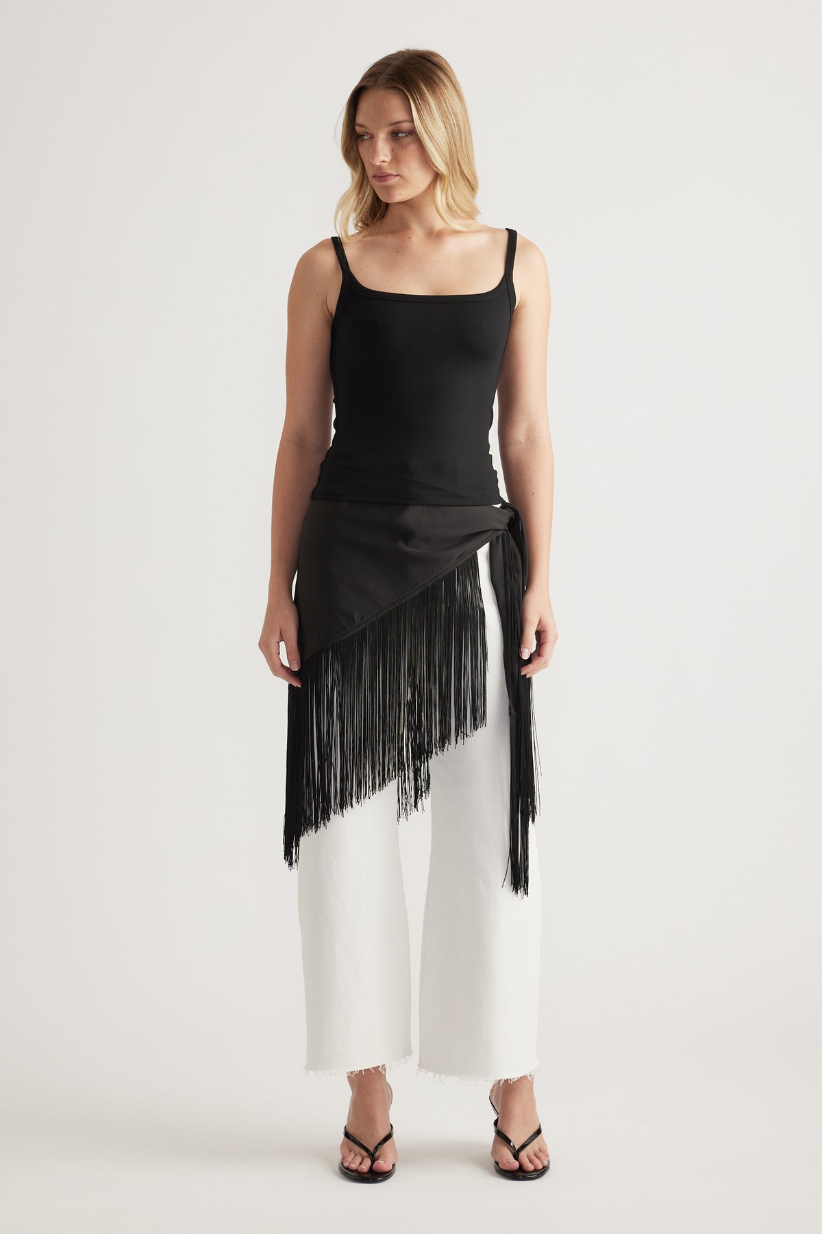 Woman wearing a black thin strap fitted tank with a fringed wrap overlay, white wide leg pants and sandals