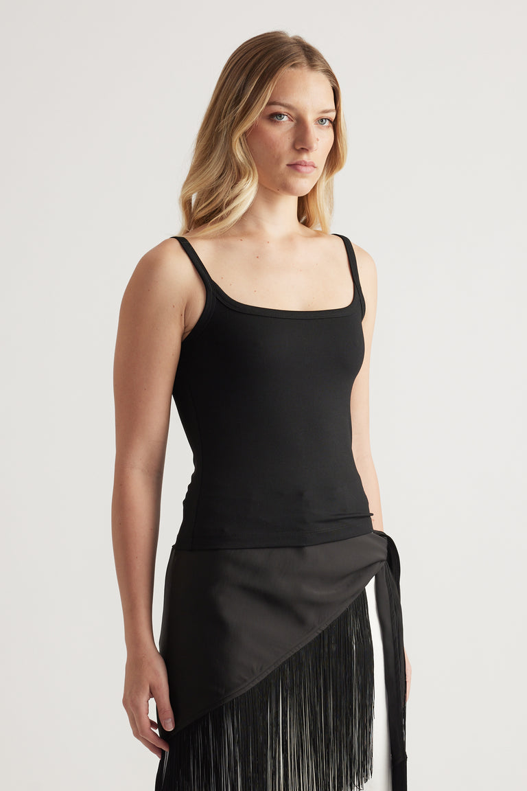 Woman wearing a fitted black thin strap tank top with a square neckline and black fringed wrap skirt
