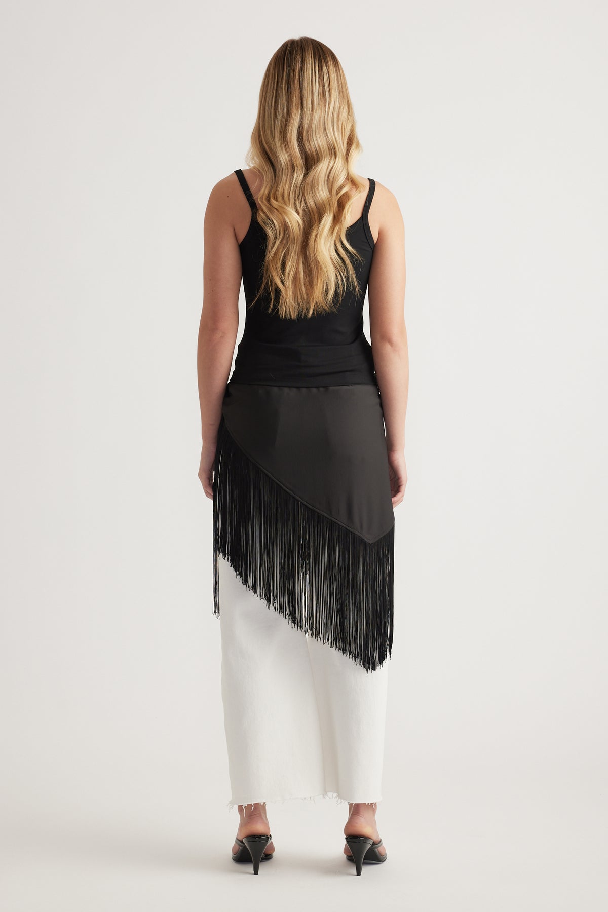 Model viewed from behind wearing a fitted black thin strap tank with white wide leg pants and a fringed asymmetric overlay