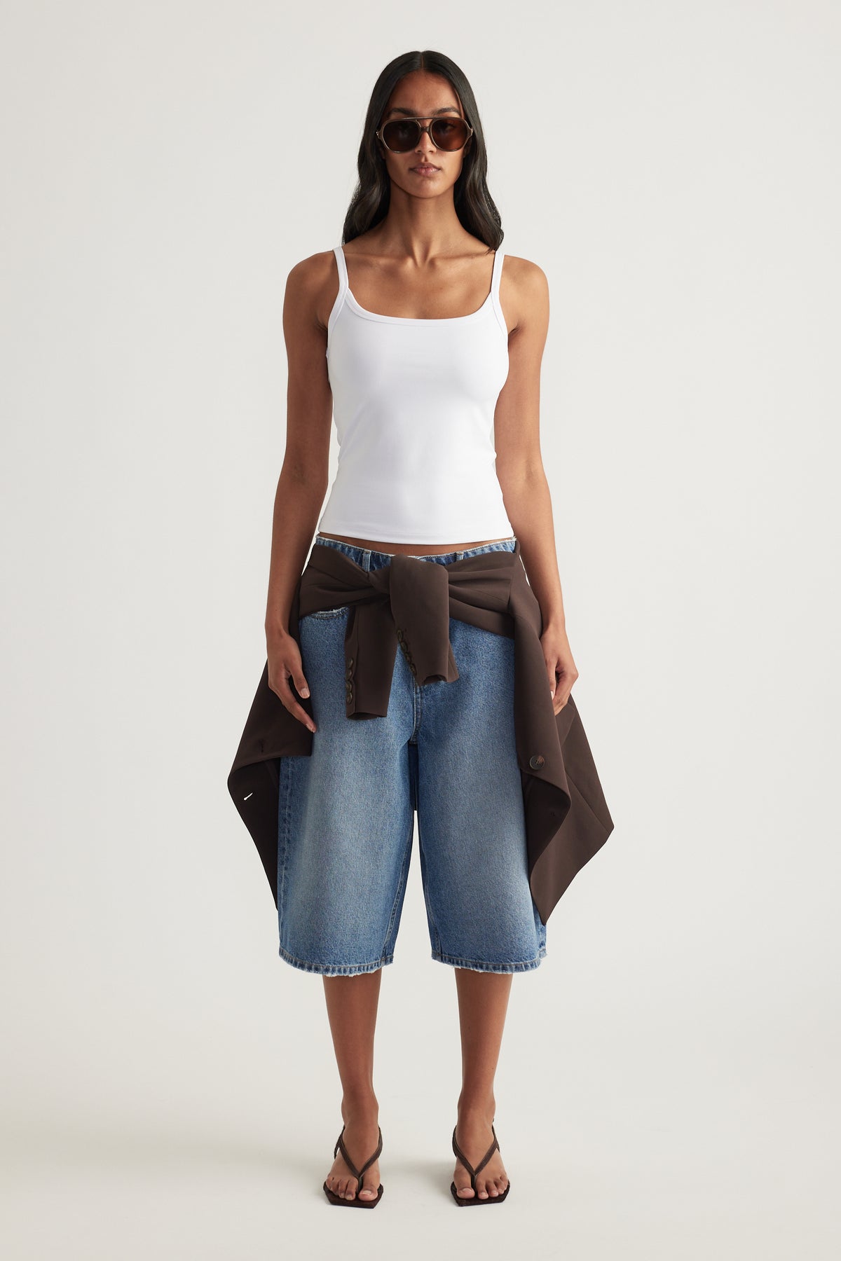 Woman wearing a white fitted thin strap tank with denim knee length shorts and a brown jacket tied at the waist
