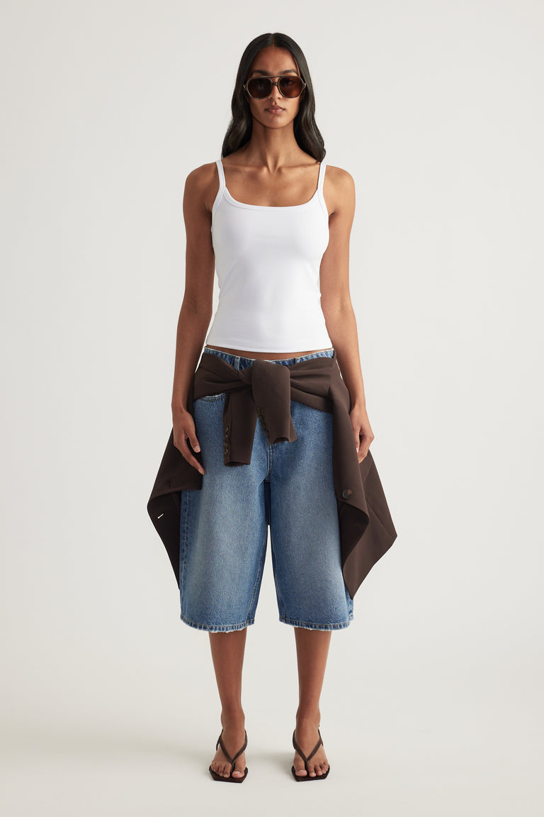 Woman wearing a white fitted thin strap tank with denim knee length shorts and a brown jacket tied at the waist