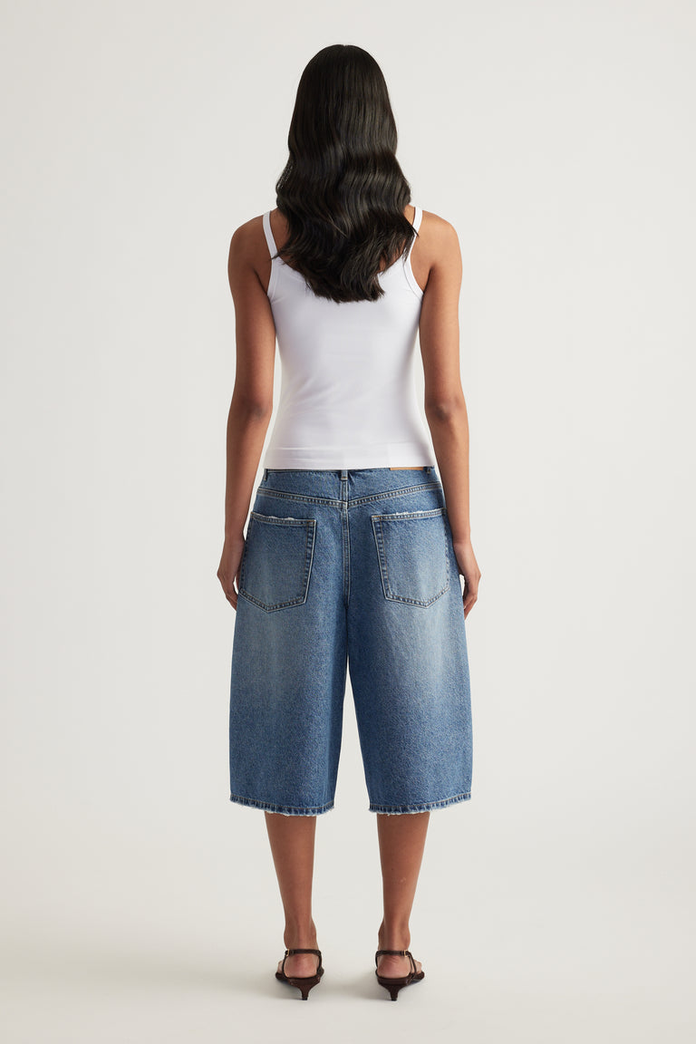 Model viewed from behind wearing a white fitted thin strap tank with blue denim knee-length shorts