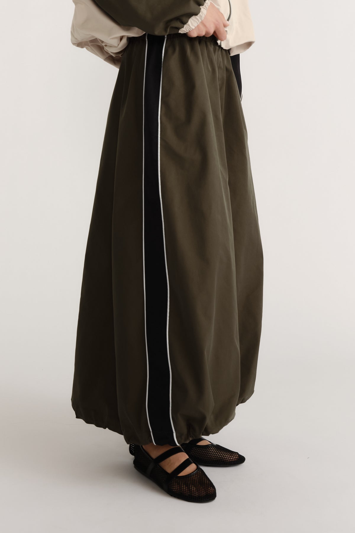 Side view of ankle-length olive skirt with cocoon shape and contrasting vertical panel detail worn with black mesh flats