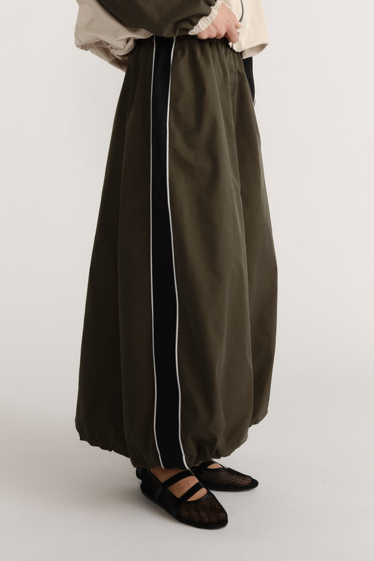 Side view of ankle-length olive skirt with cocoon shape and contrasting vertical panel detail worn with black mesh flats