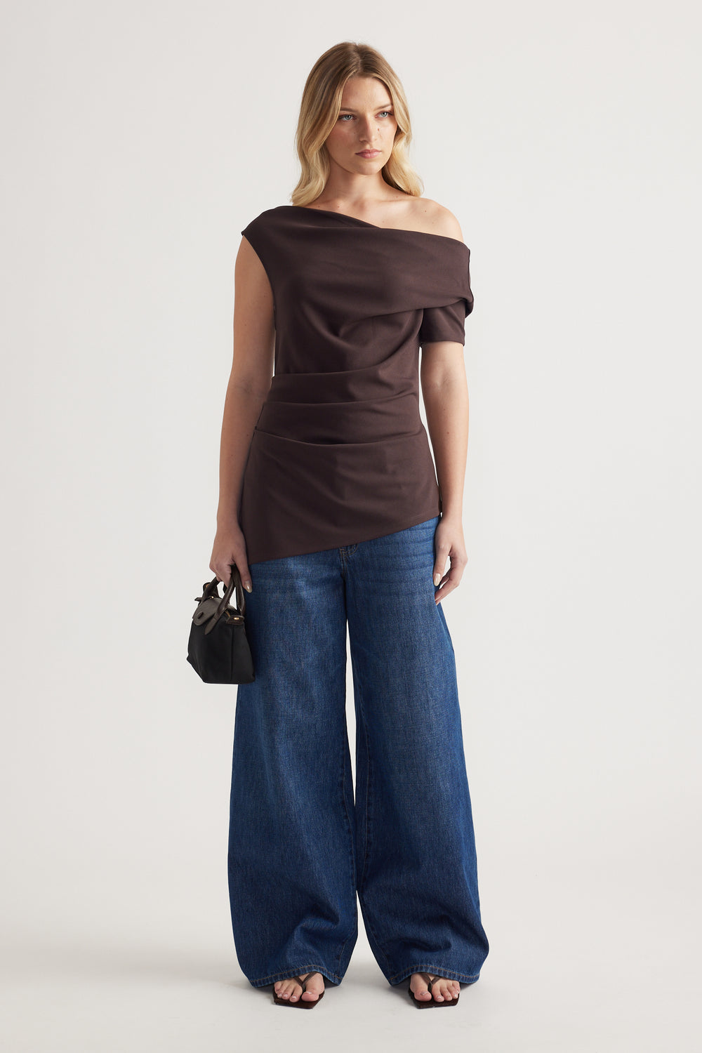 Woman wearing a dark brown off shoulder asymmetric top with blue wide leg jeans and a black handbag