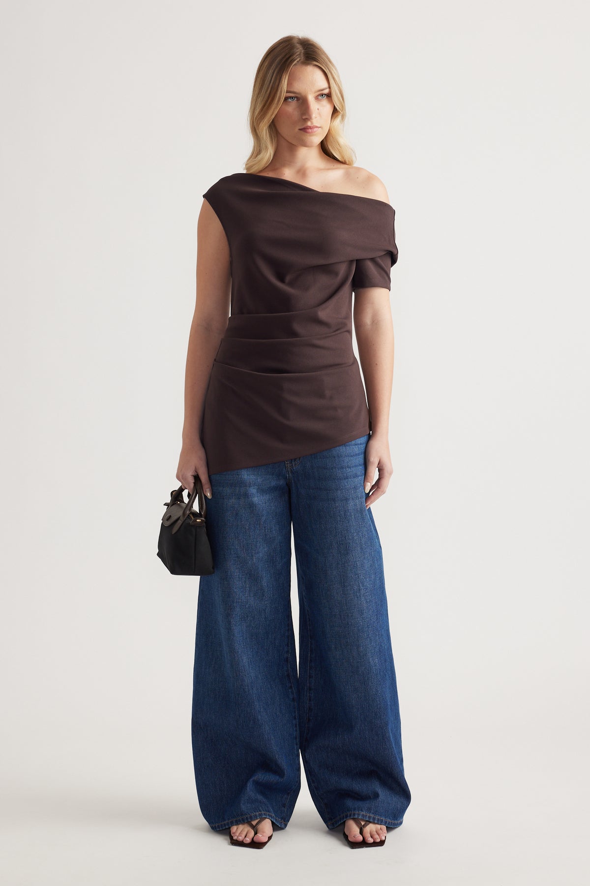Woman wearing a dark brown off shoulder asymmetric top with blue wide leg jeans and a black handbag