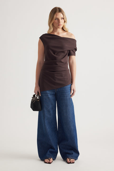 Woman wearing a dark brown off shoulder asymmetric top with blue wide leg jeans and a black handbag