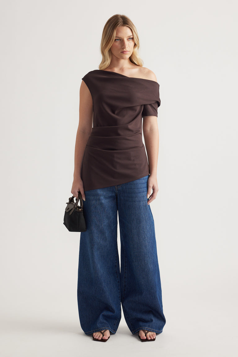 Woman wearing a dark brown off shoulder asymmetric top with blue wide leg jeans and a black handbag