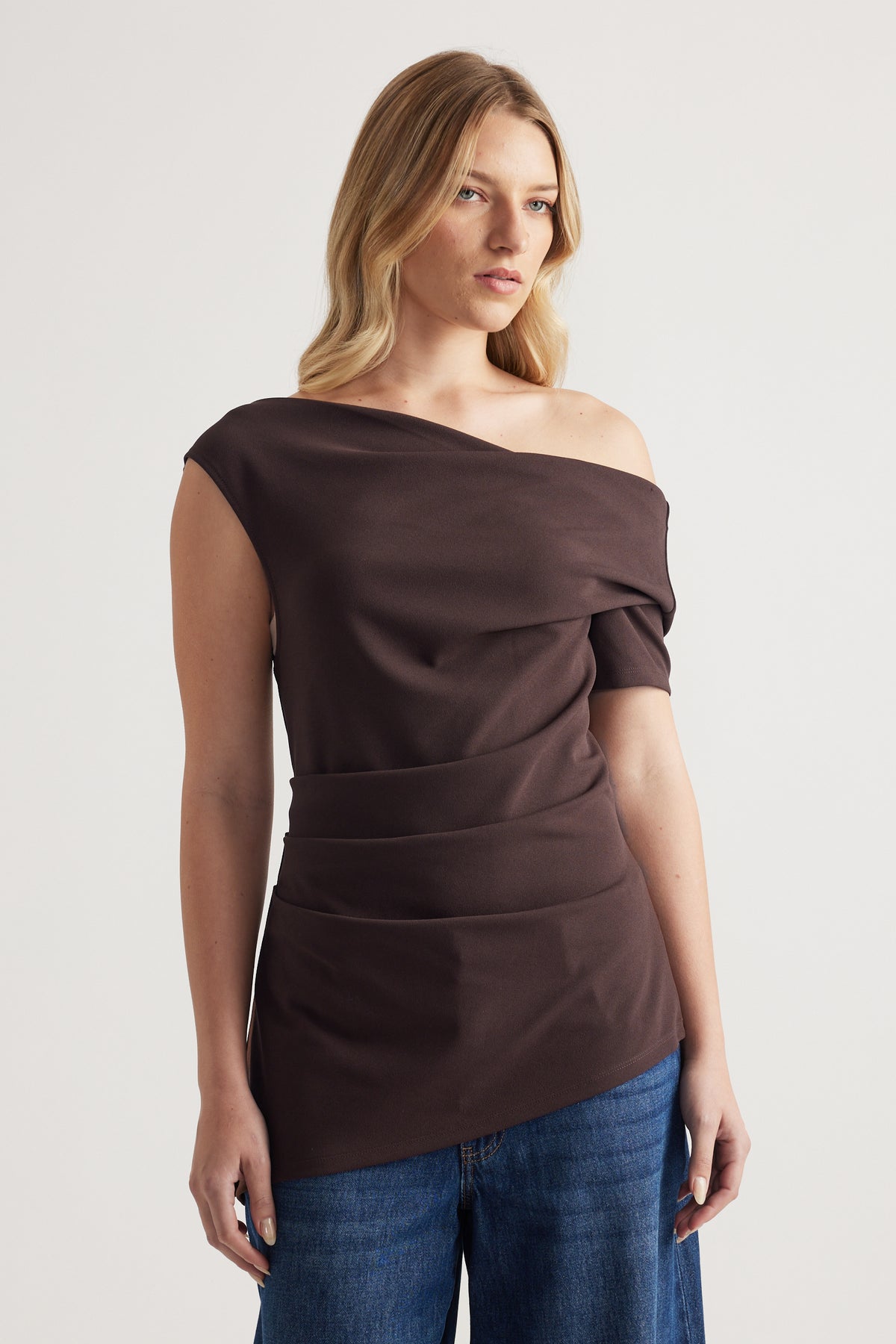 Woman wearing a dark brown off shoulder asymmetric top with crossover detailing and blue jeans