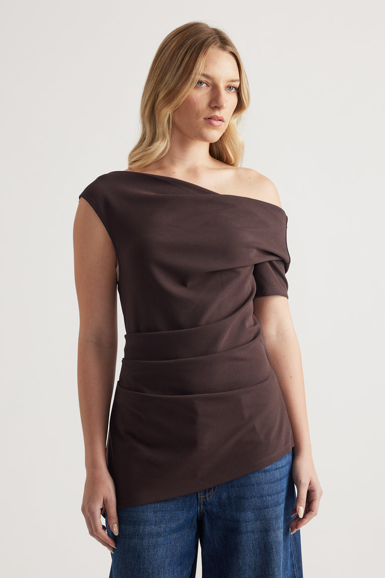 Woman wearing a dark brown off shoulder asymmetric top with crossover detailing and blue jeans