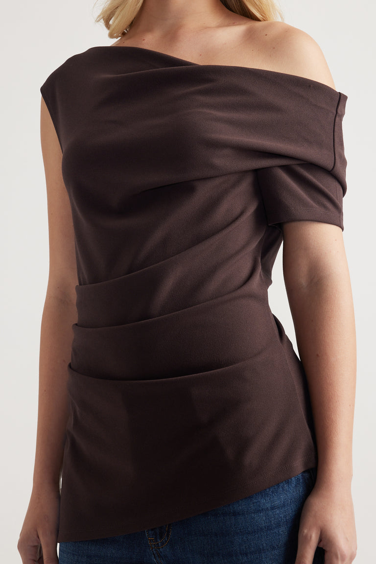 Woman wearing a dark brown off shoulder top with crossover drape and asymmetric hem over blue jeans