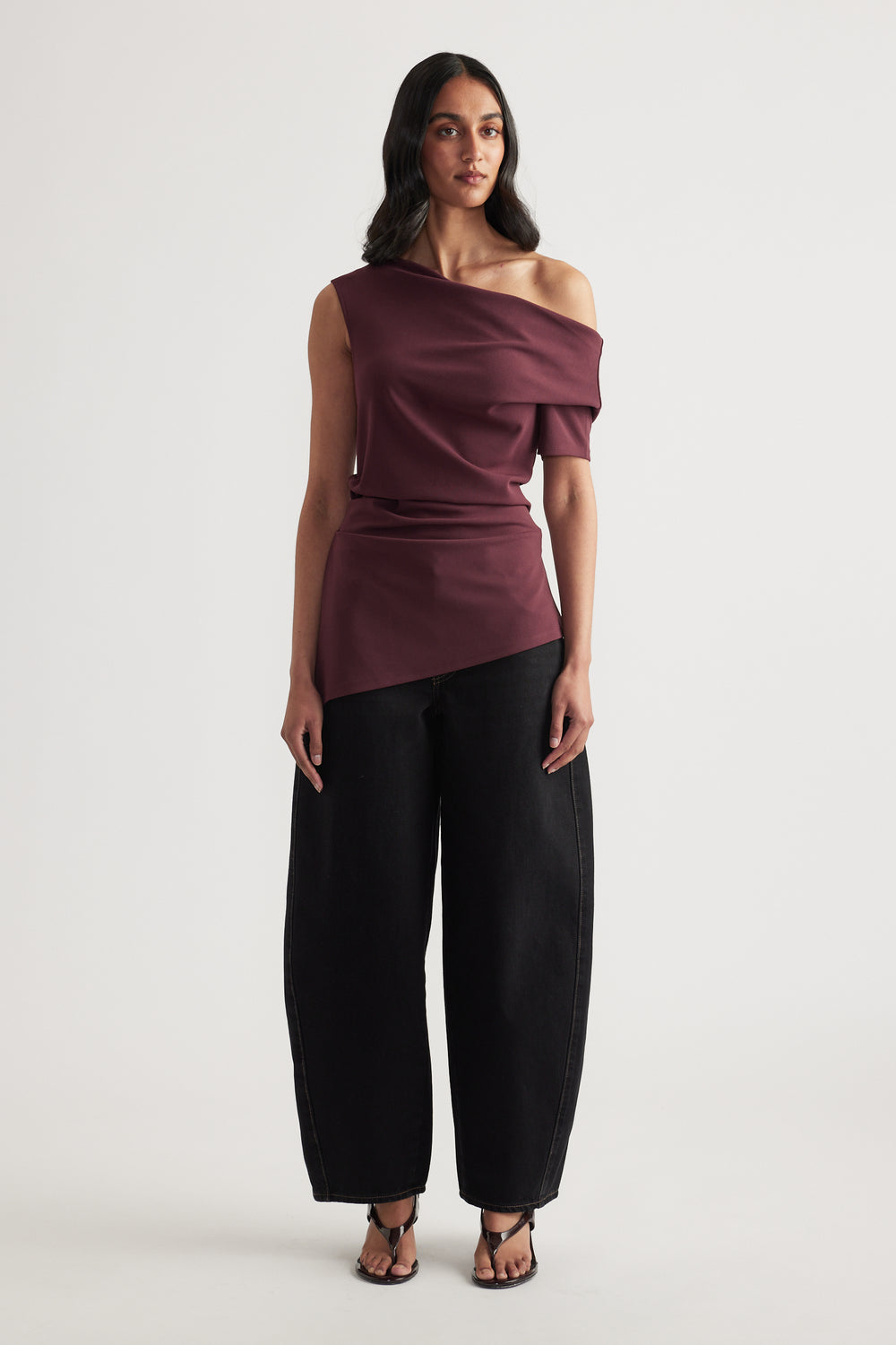 Woman wearing a burgundy off shoulder asymmetric top with crossover detail, black wide leg pants and sandals
