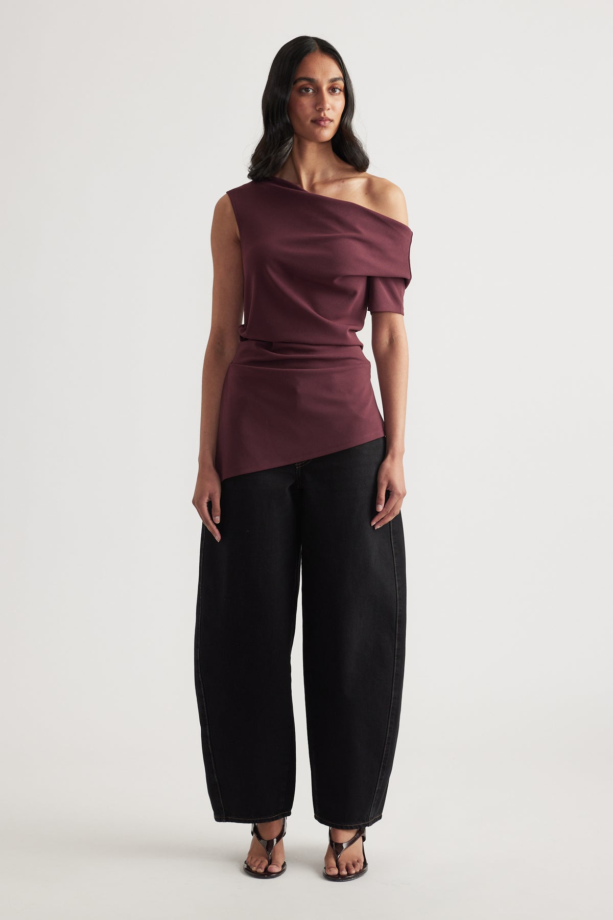 Woman wearing a burgundy off shoulder asymmetric top with crossover detail, black wide leg pants and sandals