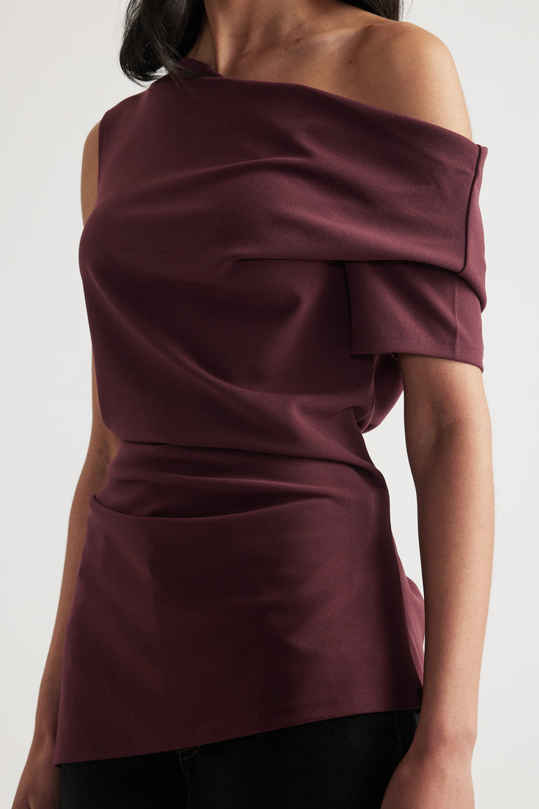 Woman wearing a burgundy off shoulder top with crossover draping and asymmetric hem with black pants