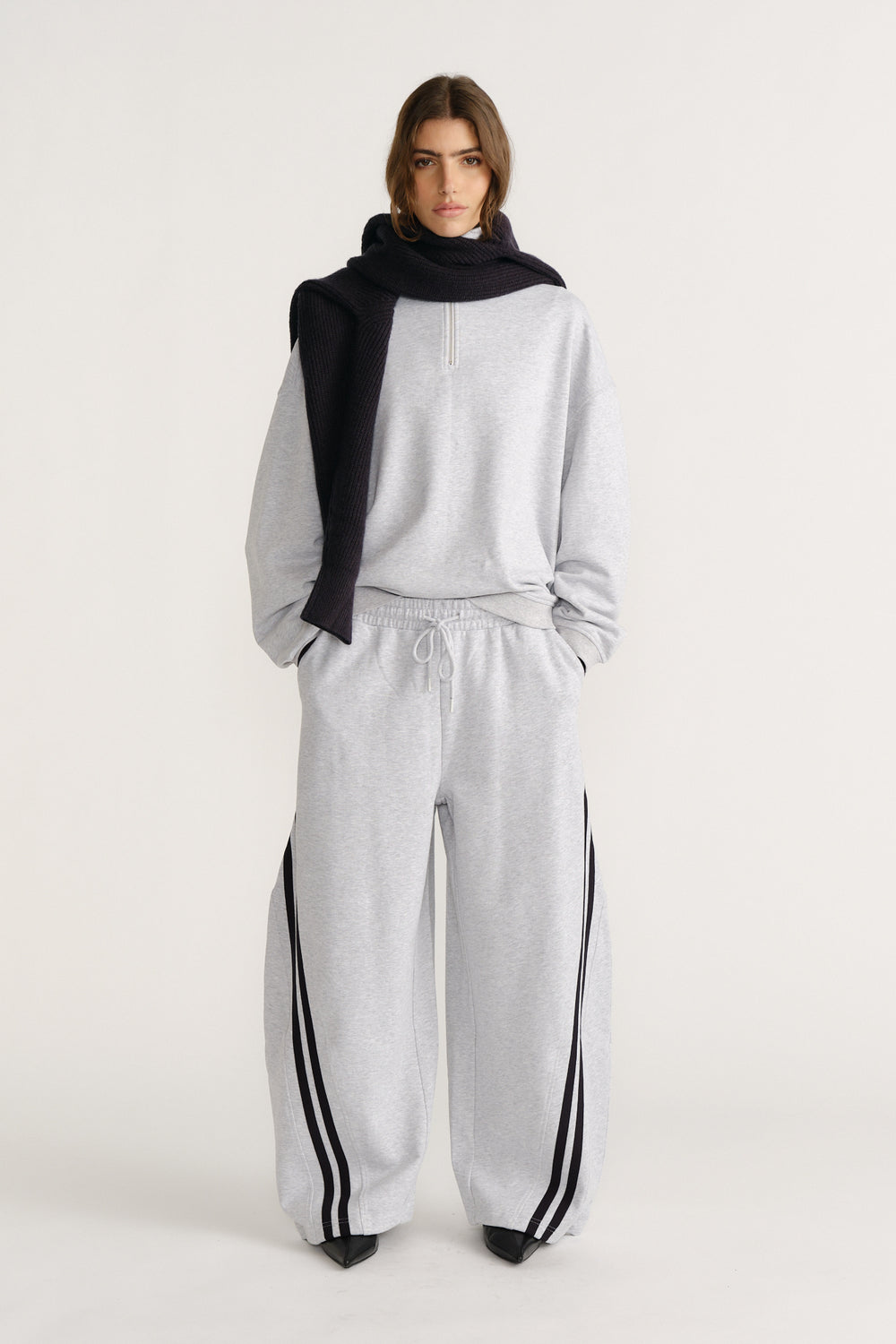 Woman wearing relaxed light grey trackpants with black side stripes, paired with a matching pullover and black knit scarf draped over shoulders
