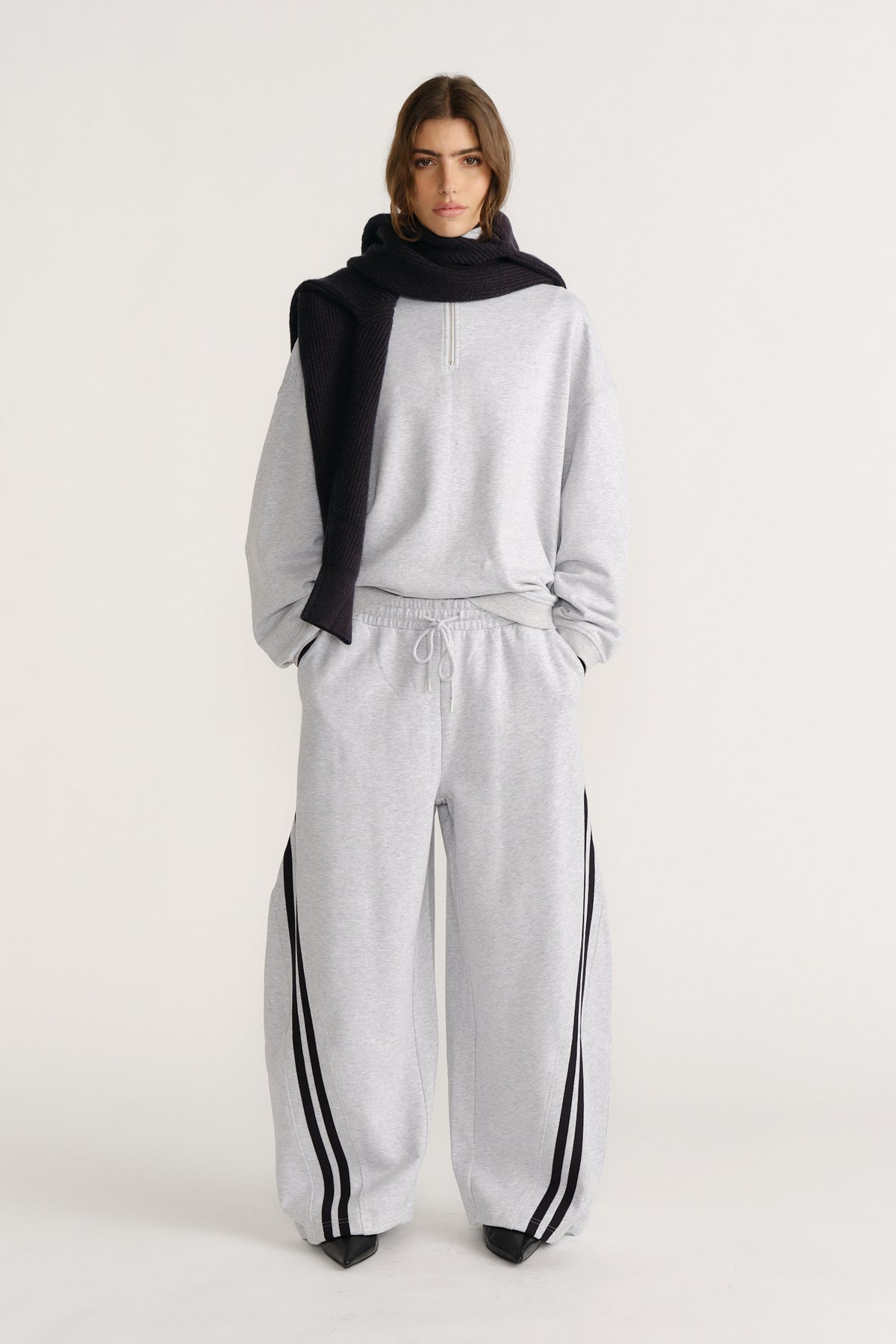 Woman wearing relaxed light grey trackpants with black side stripes, paired with a matching pullover and black knit scarf draped over shoulders