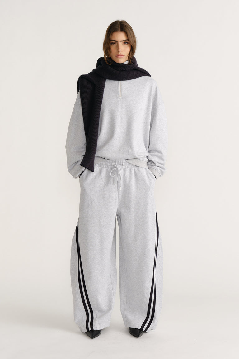 Woman wearing relaxed light grey trackpants with black side stripes, paired with a matching pullover and black knit scarf draped over shoulders
