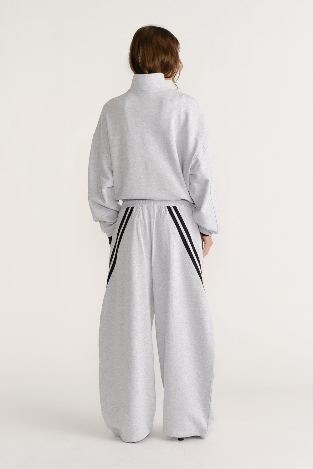 Woman seen from behind in light grey wide-leg trackpants with black stripe details and a matching high-neck sweatshirt
