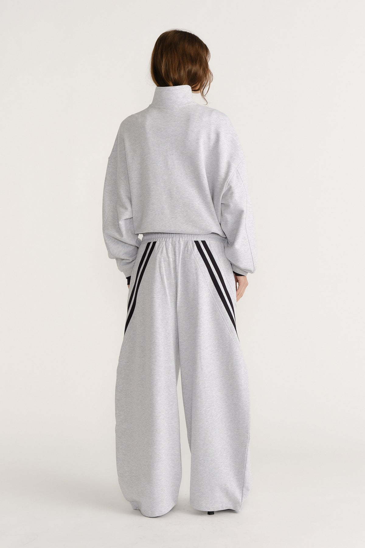Woman seen from behind in light grey wide-leg trackpants with black stripe details and a matching high-neck sweatshirt