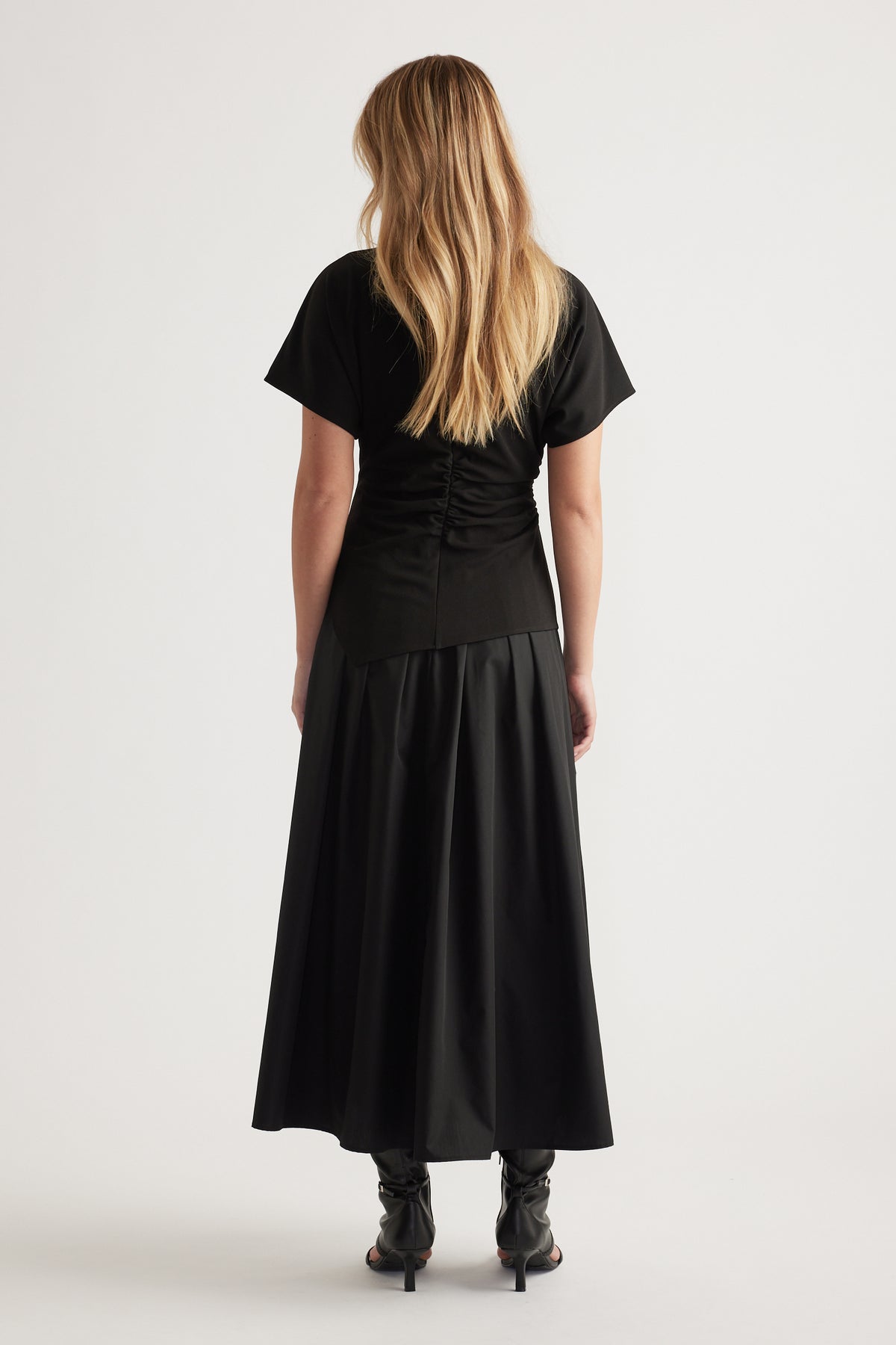 Back view of woman wearing a black spliced midi dress with short sleeves and relaxed silhouette, styled with black heeled boots
