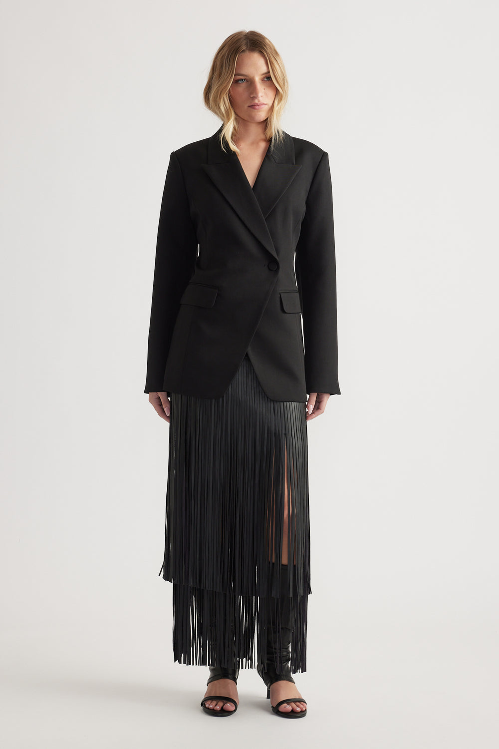 Woman wearing a black tailored blazer with padded shoulders paired with a long black pleated skirt with a front slit