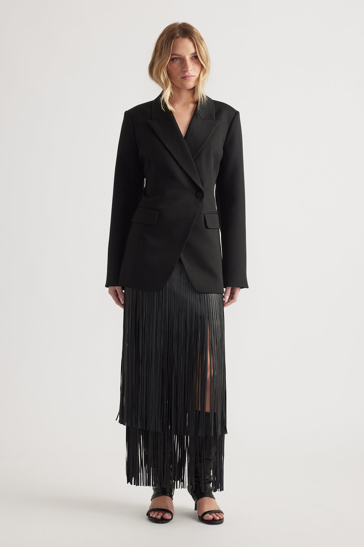 Woman wearing a black tailored blazer with padded shoulders paired with a long black pleated skirt with a front slit