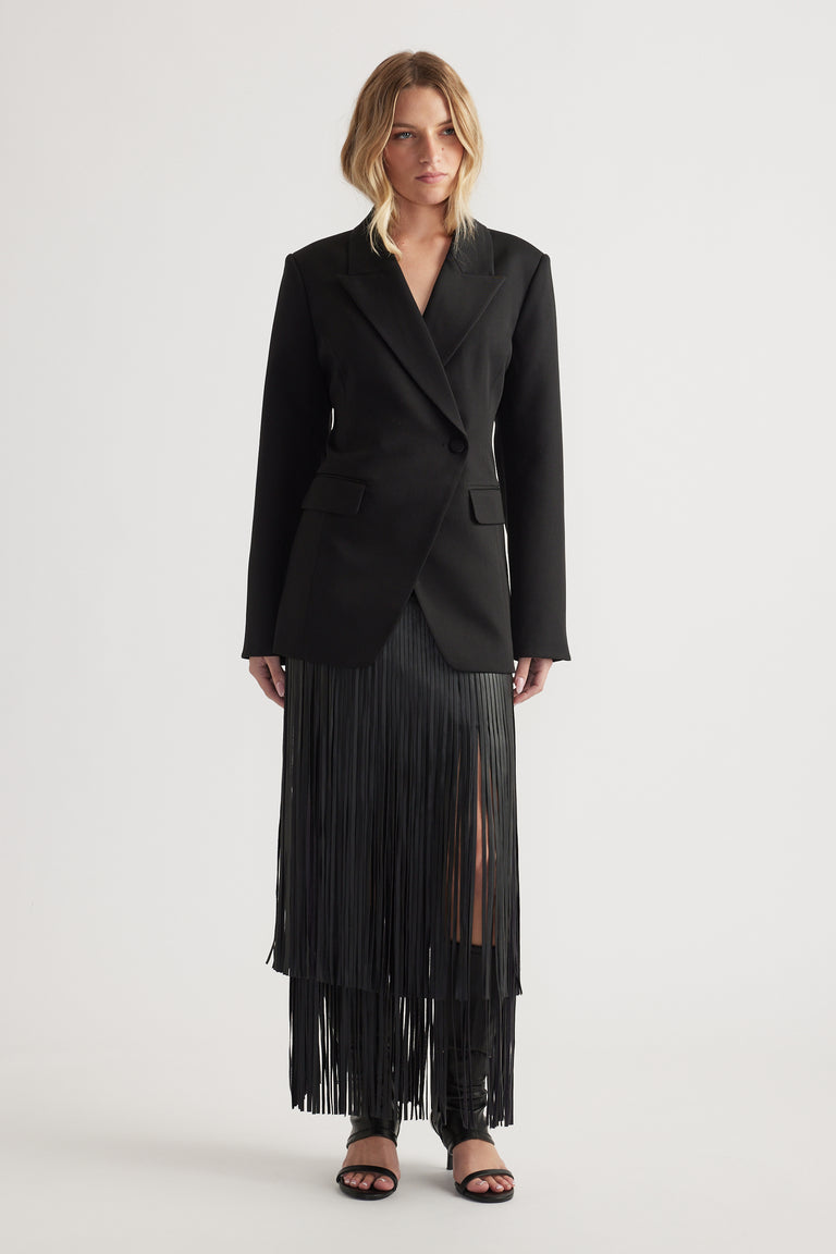 Woman wearing a black tailored blazer with padded shoulders paired with a long black pleated skirt with a front slit