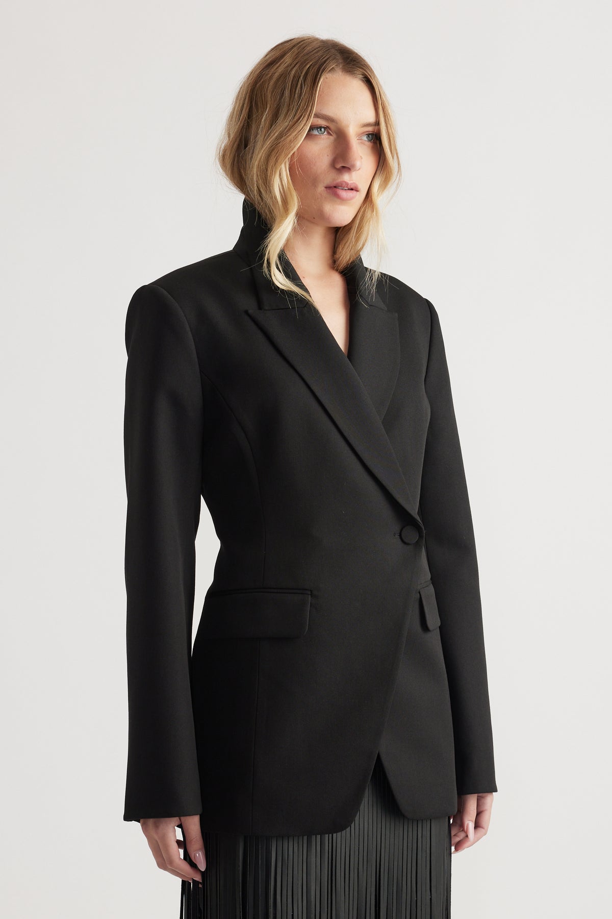 Black tailored blazer with structured shoulders, asymmetric button front, collar, and flap pockets on model