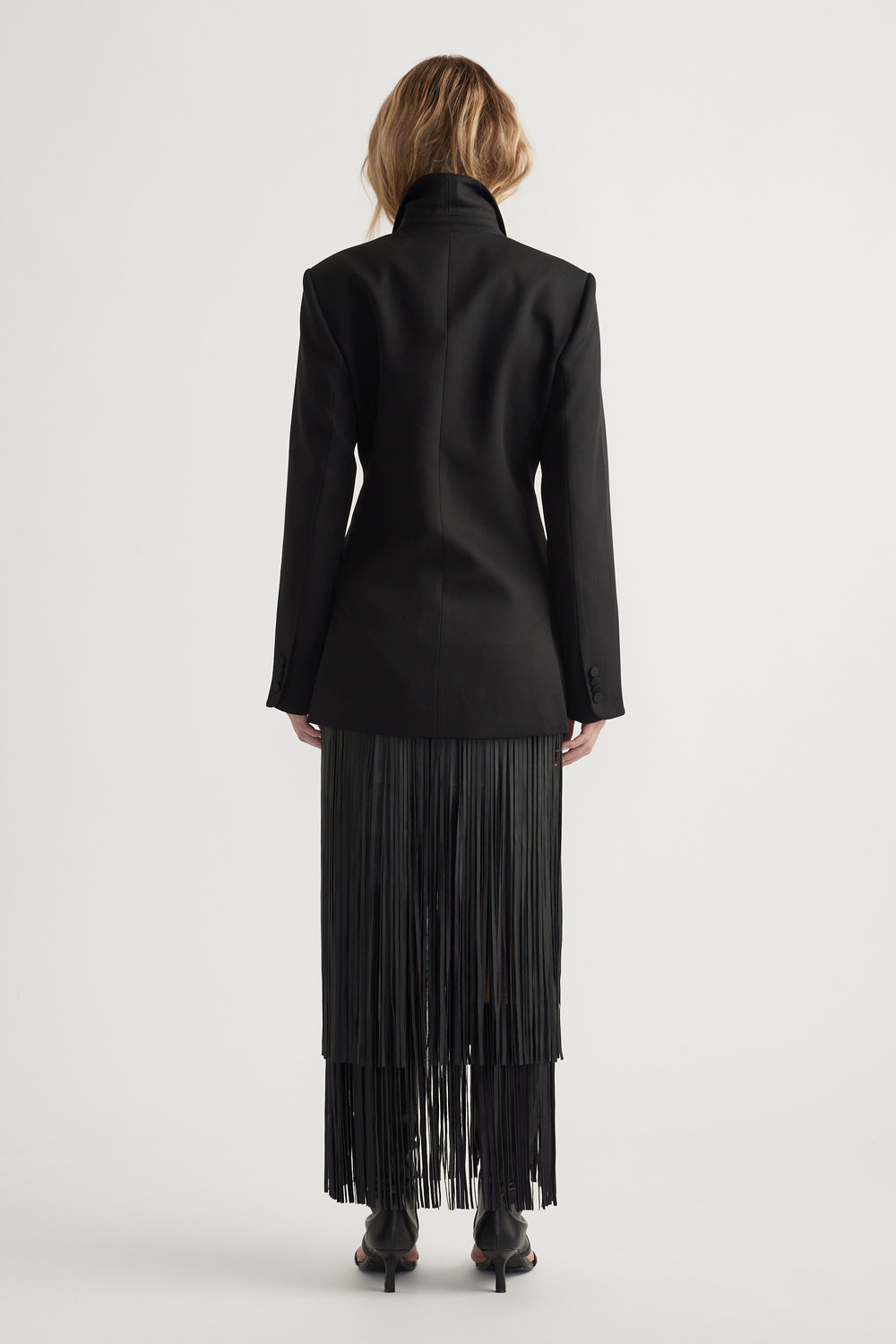 Back view of a fitted black blazer with padded shoulders paired with a fringed black skirt and black heels on model