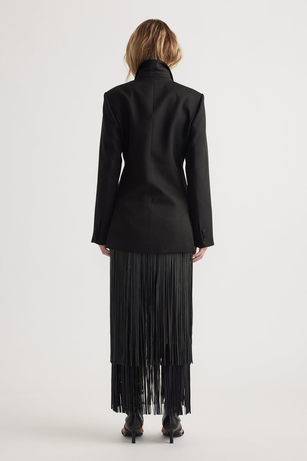 Back view of a fitted black blazer with padded shoulders paired with a fringed black skirt and black heels on model