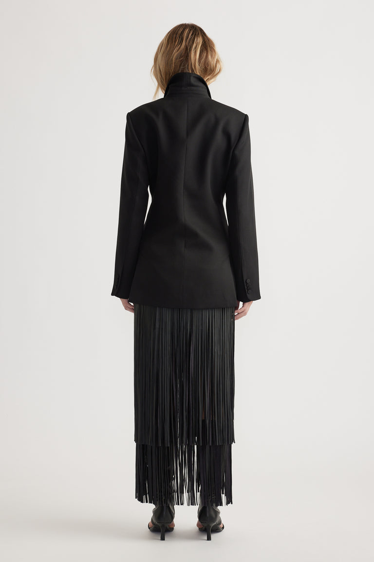 Back view of a fitted black blazer with padded shoulders paired with a fringed black skirt and black heels on model