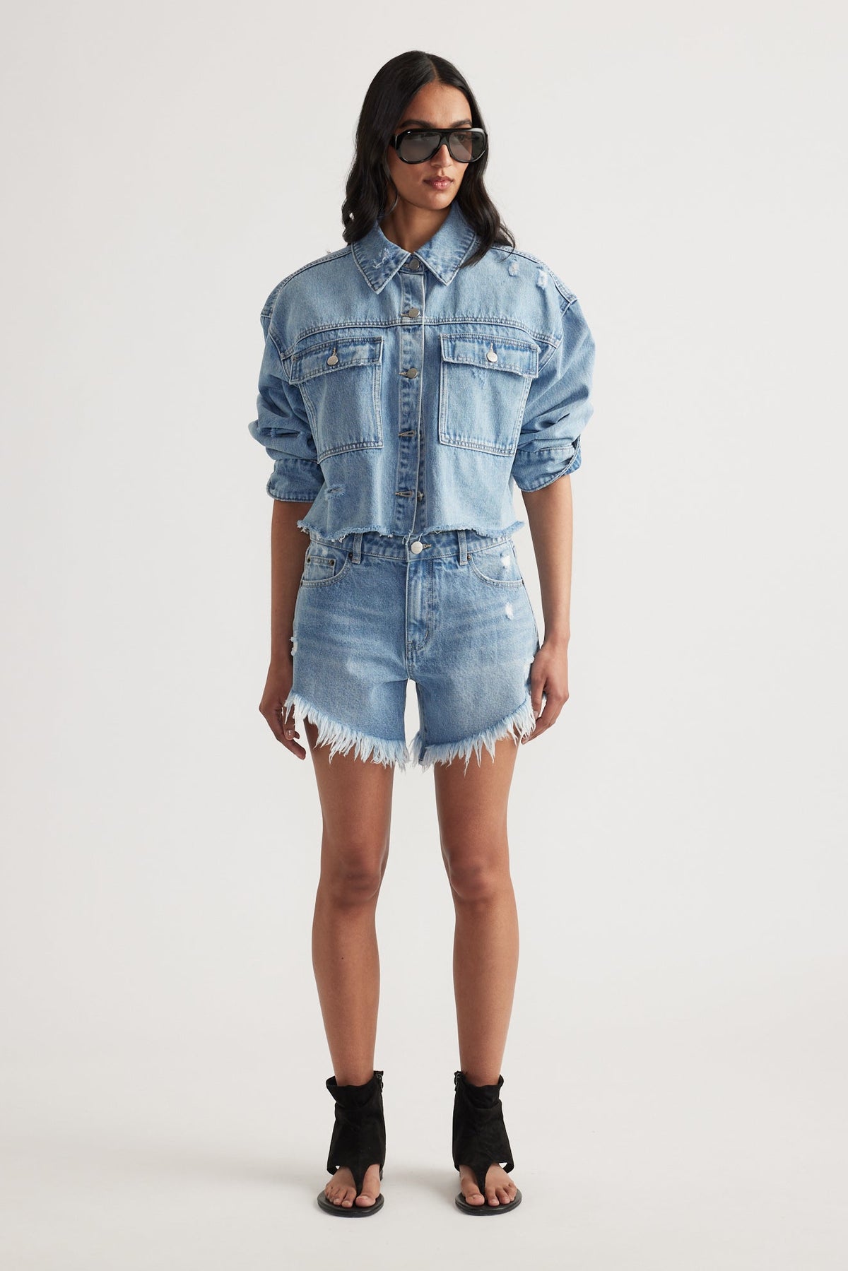 Woman wearing a cropped denim jacket paired with high rise, frayed hem distressed denim shorts and black open-toe heels