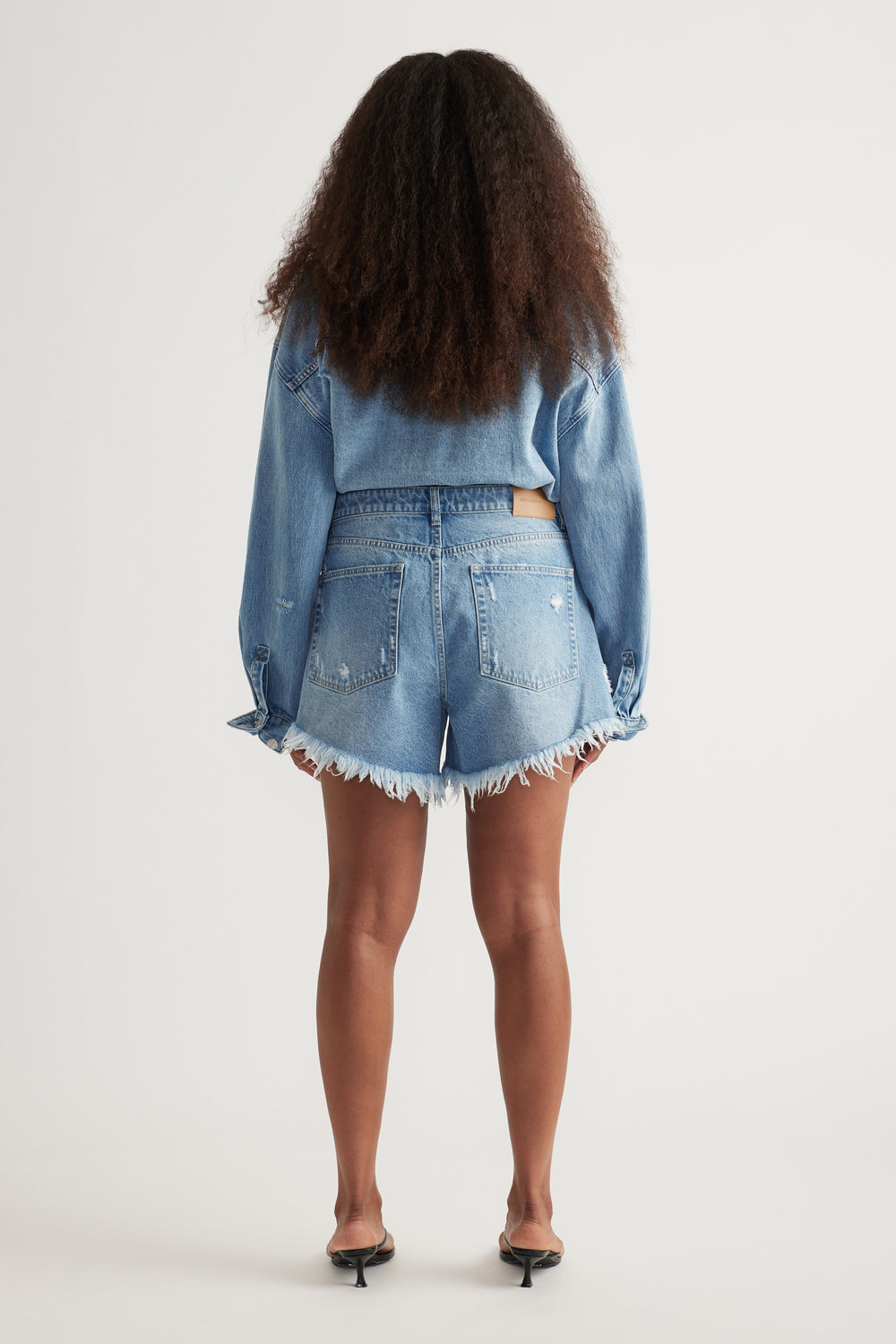 Woman wearing high rise distressed denim shorts with frayed hems paired with a loose light denim jacket
