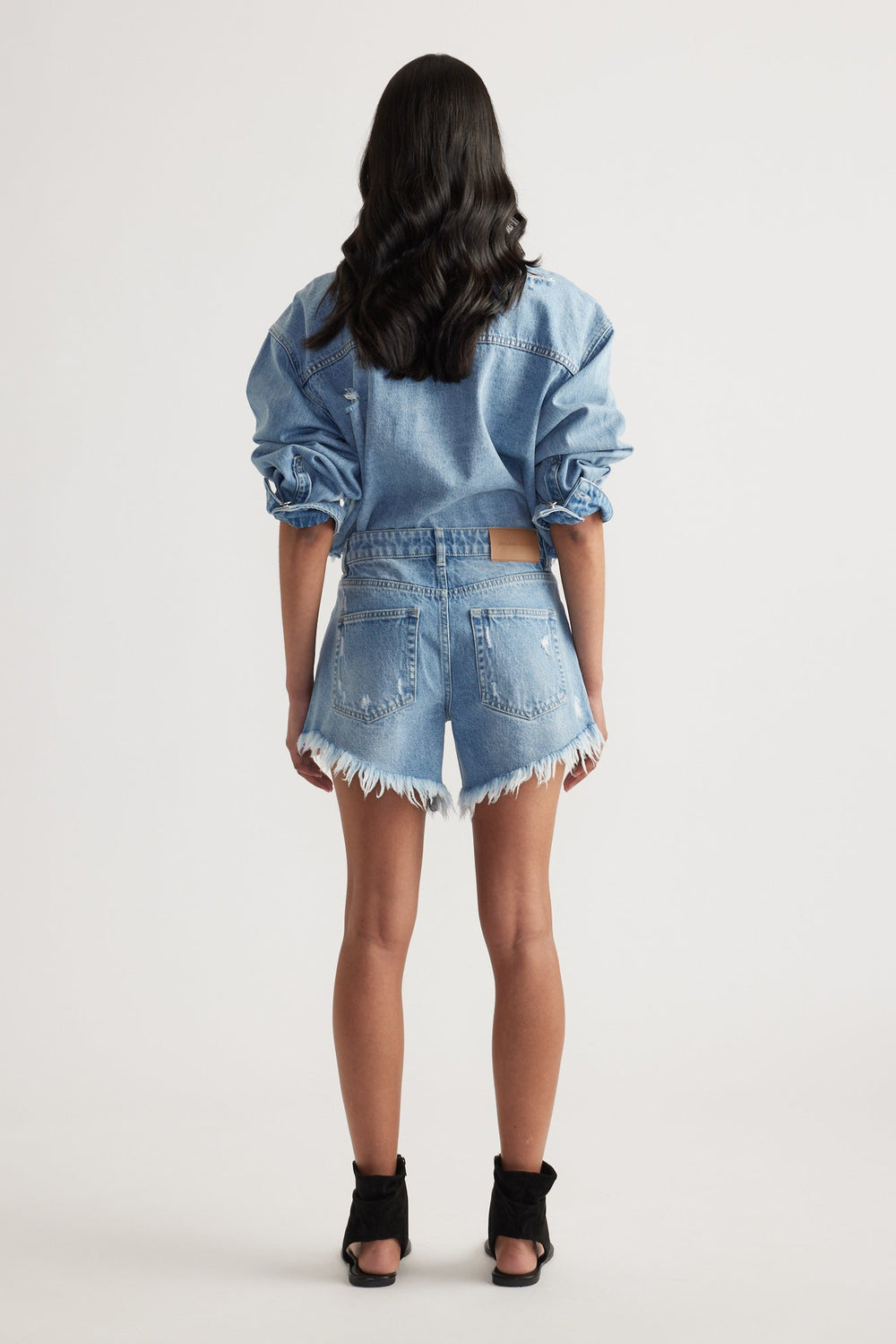 Rear view of a woman wearing high-rise light blue denim shorts with distressed frayed hems and a matching denim jacket