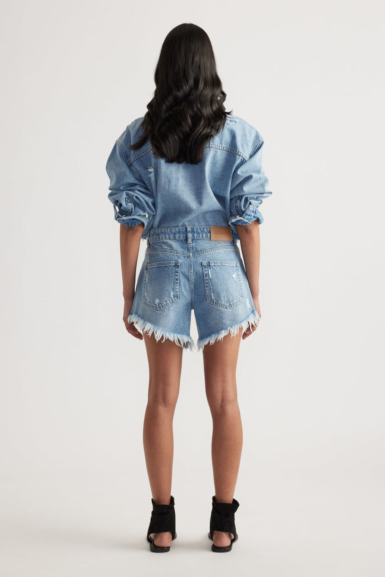 Rear view of a woman wearing high-rise light blue denim shorts with distressed frayed hems and a matching denim jacket