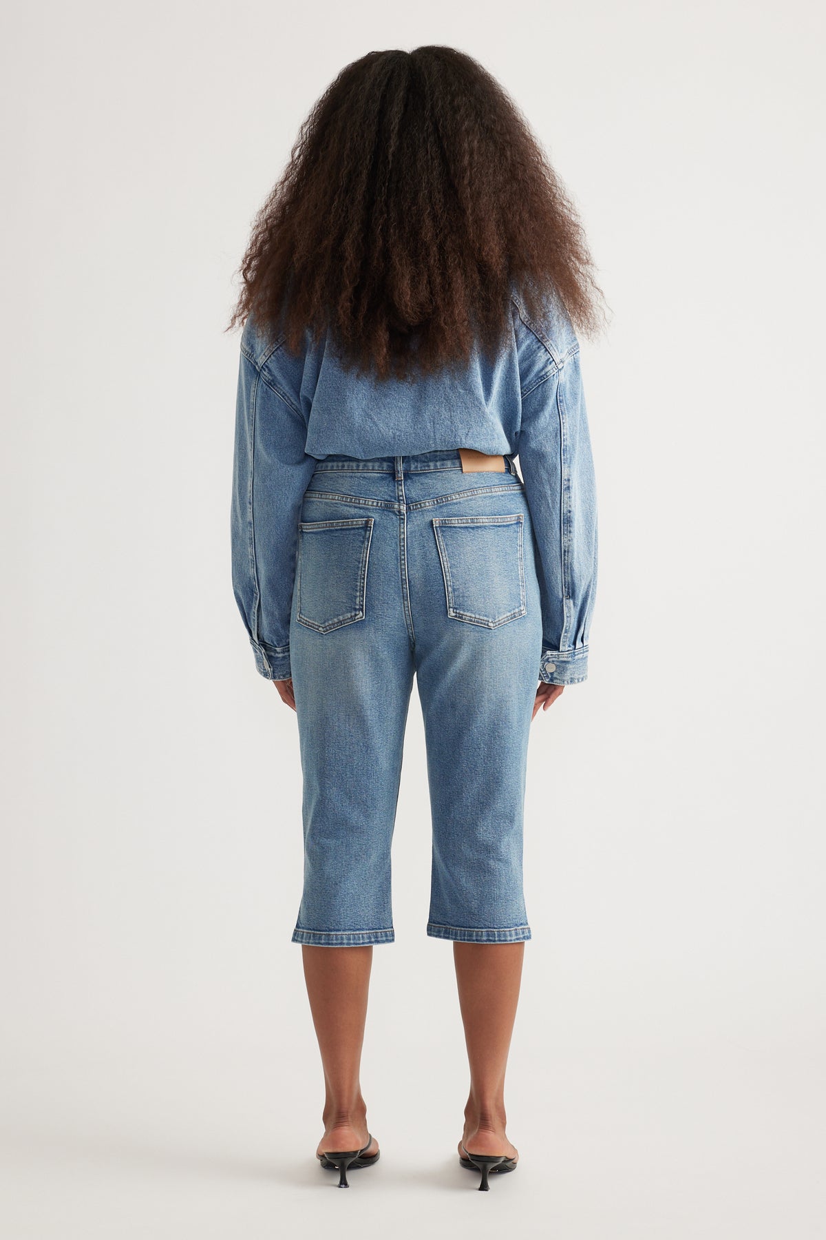 Back view of a woman wearing light wash denim capris with a matching denim shirt and black kitten heels