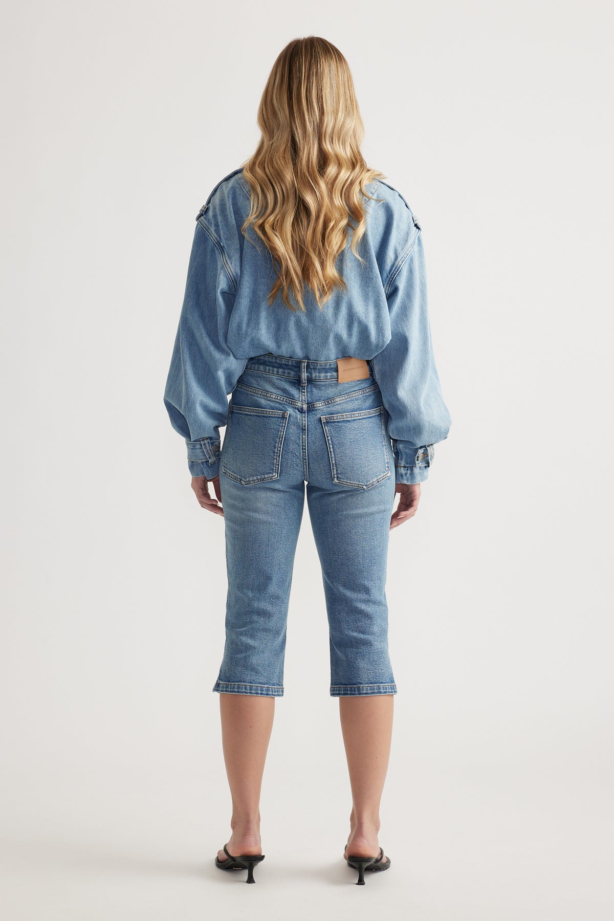 Back view of woman wearing blue high rise denim capri pants with a denim jacket and black heels