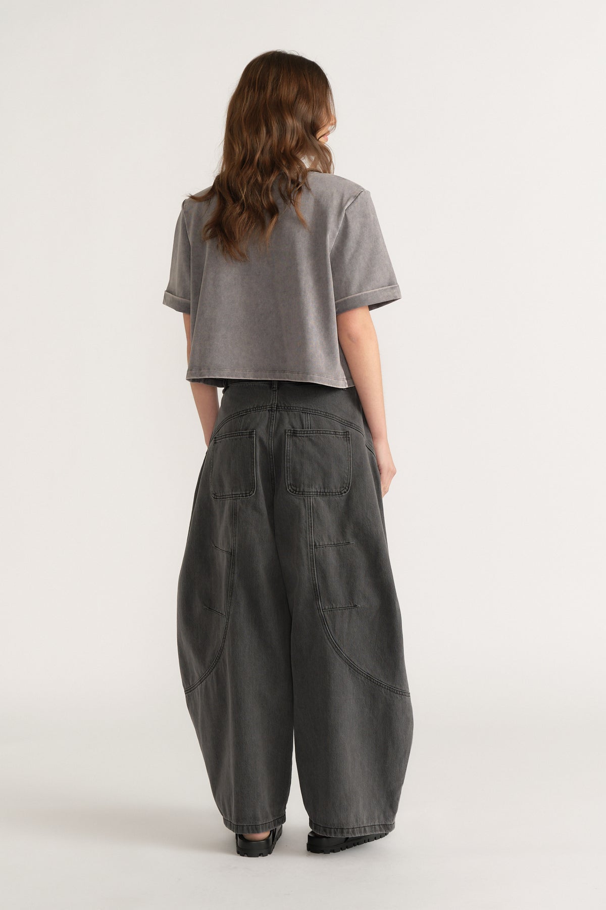 Rear view of loose-fitting dark grey denim jeans with patch pockets and seam details worn with a cropped grey t-shirt