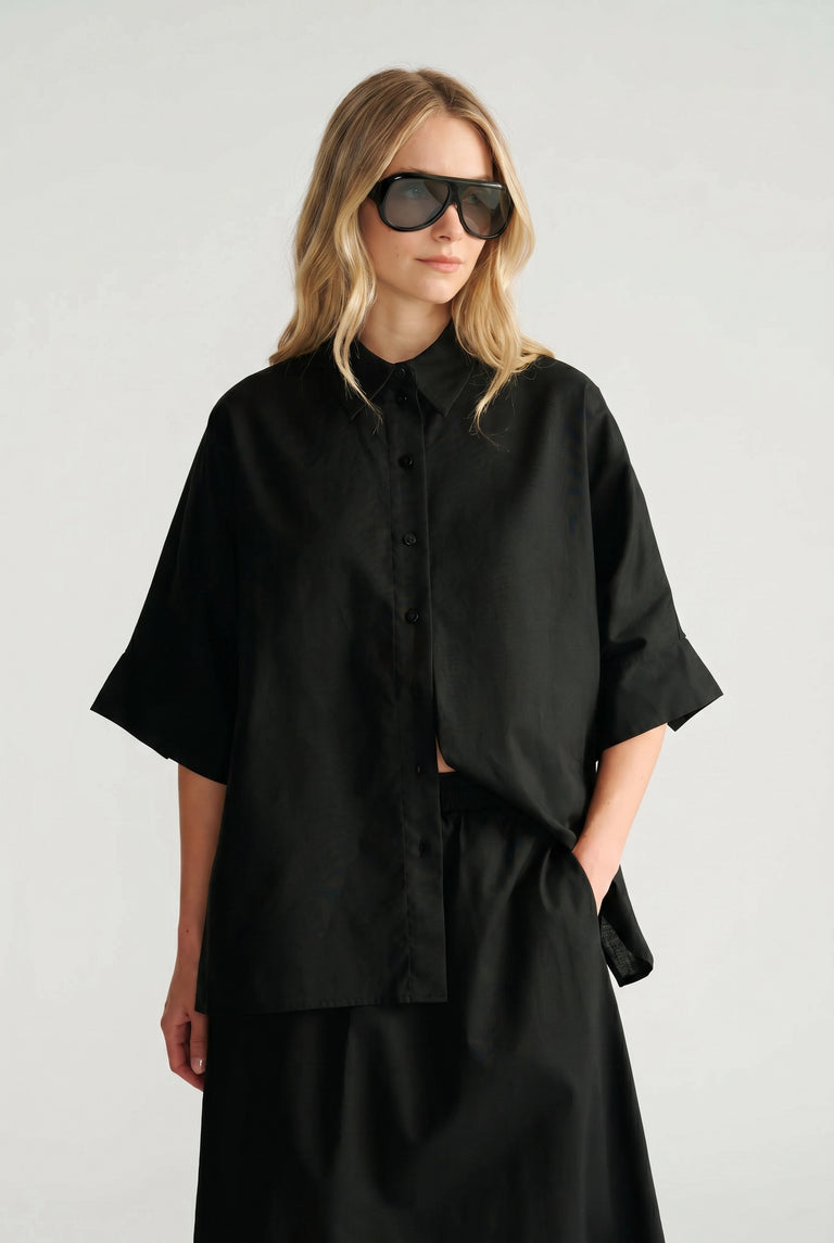 Woman wearing a relaxed black oversized shirt with wide sleeves, paired with matching black skirt and oversized sunglasses