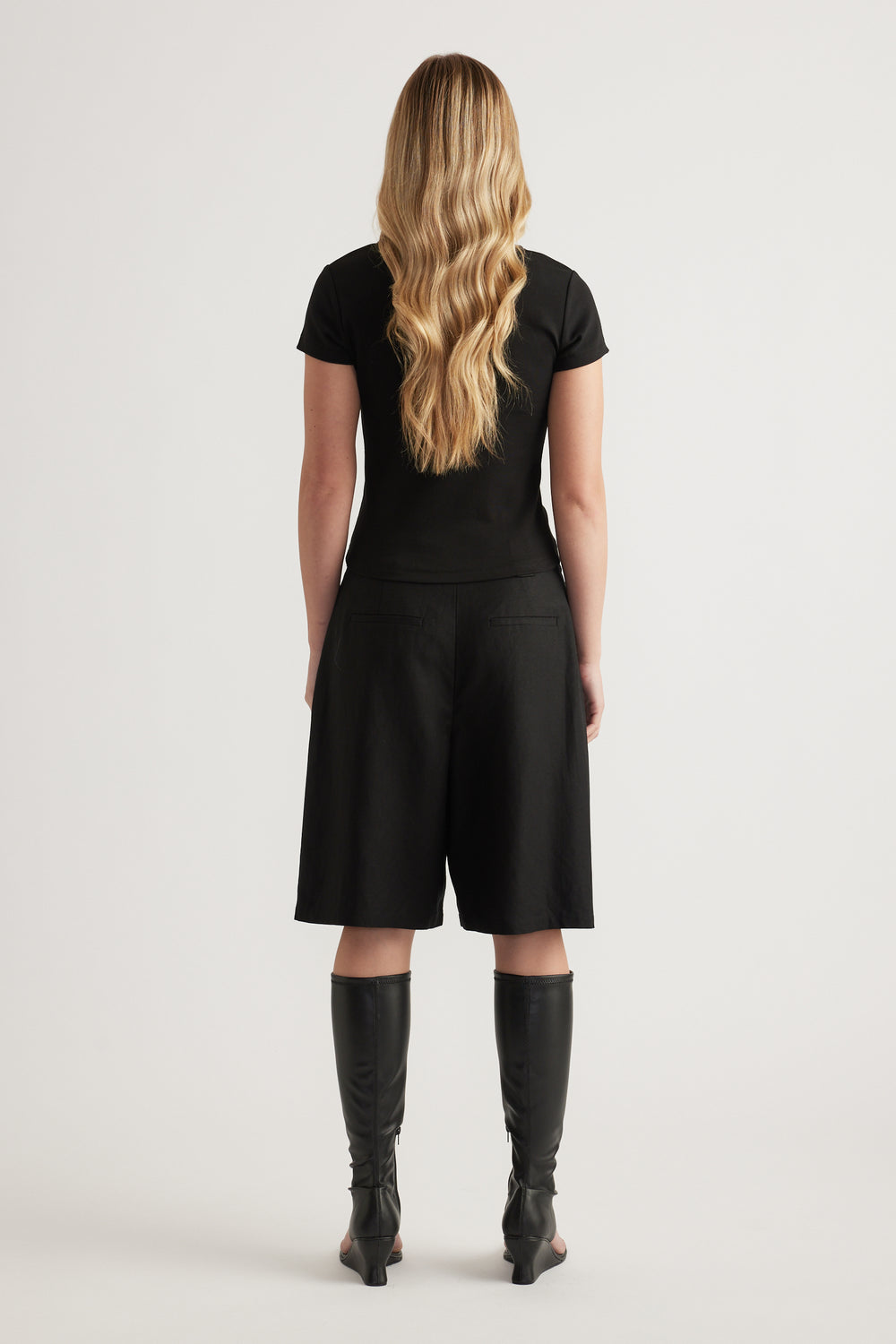 Woman viewed from behind wearing a fitted black tee with black knee length shorts and knee high boots