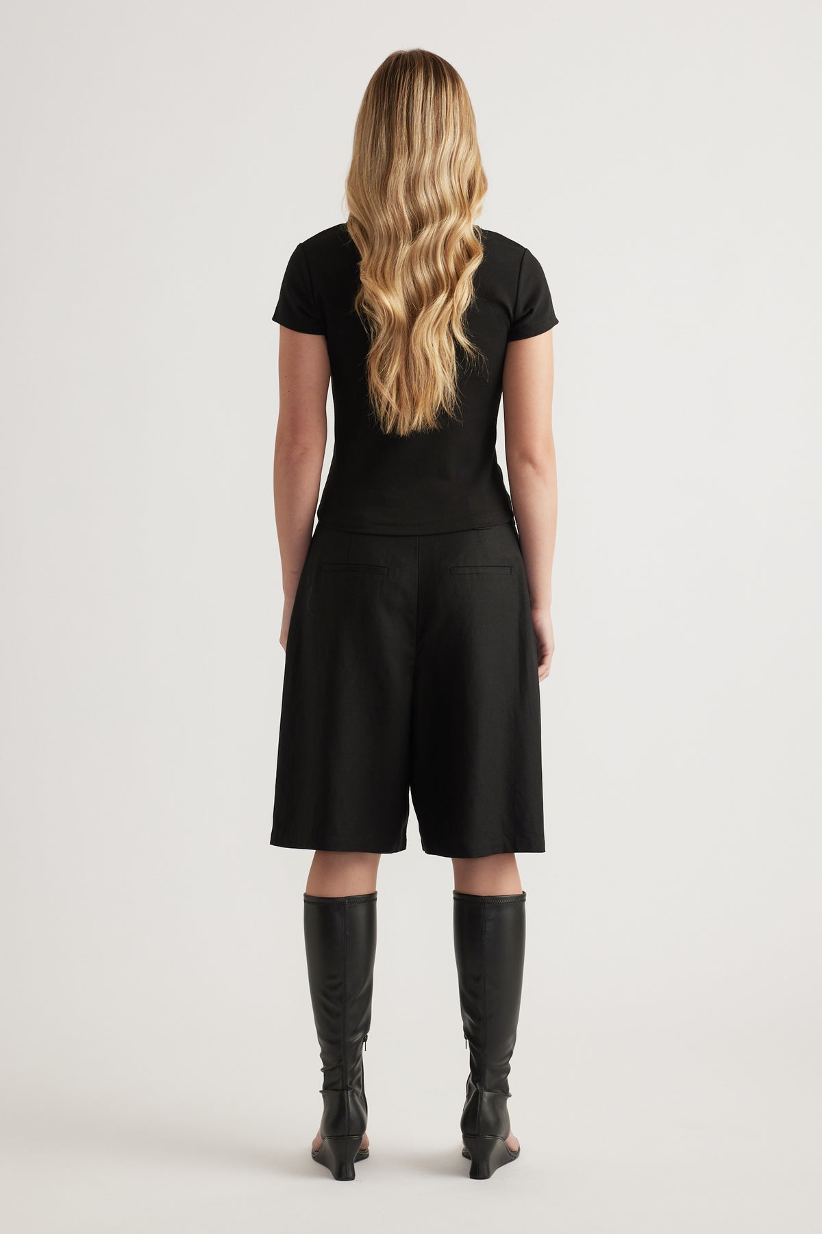 Woman viewed from behind wearing a fitted black tee with black knee length shorts and knee high boots