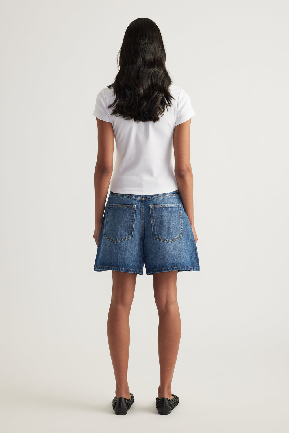 Model viewed from behind wearing a white short sleeve tee with blue denim shorts and black shoes