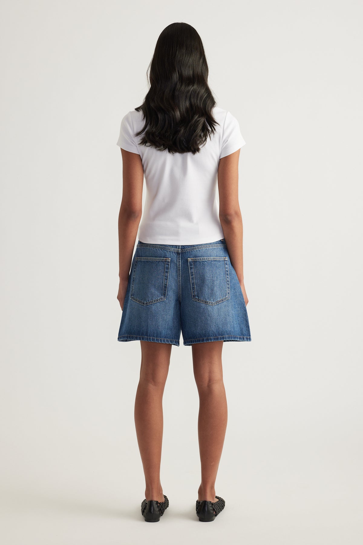 Model viewed from behind wearing a white short sleeve tee with blue denim shorts and black shoes