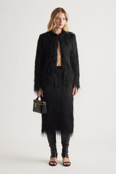 Woman wearing a textured black fringed jacket and matching skirt paired with black leather boots and a woven handbag