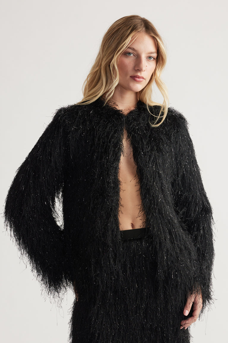 Woman wearing a black textured fringed jacket with subtle shimmer paired with matching high-waisted pants.