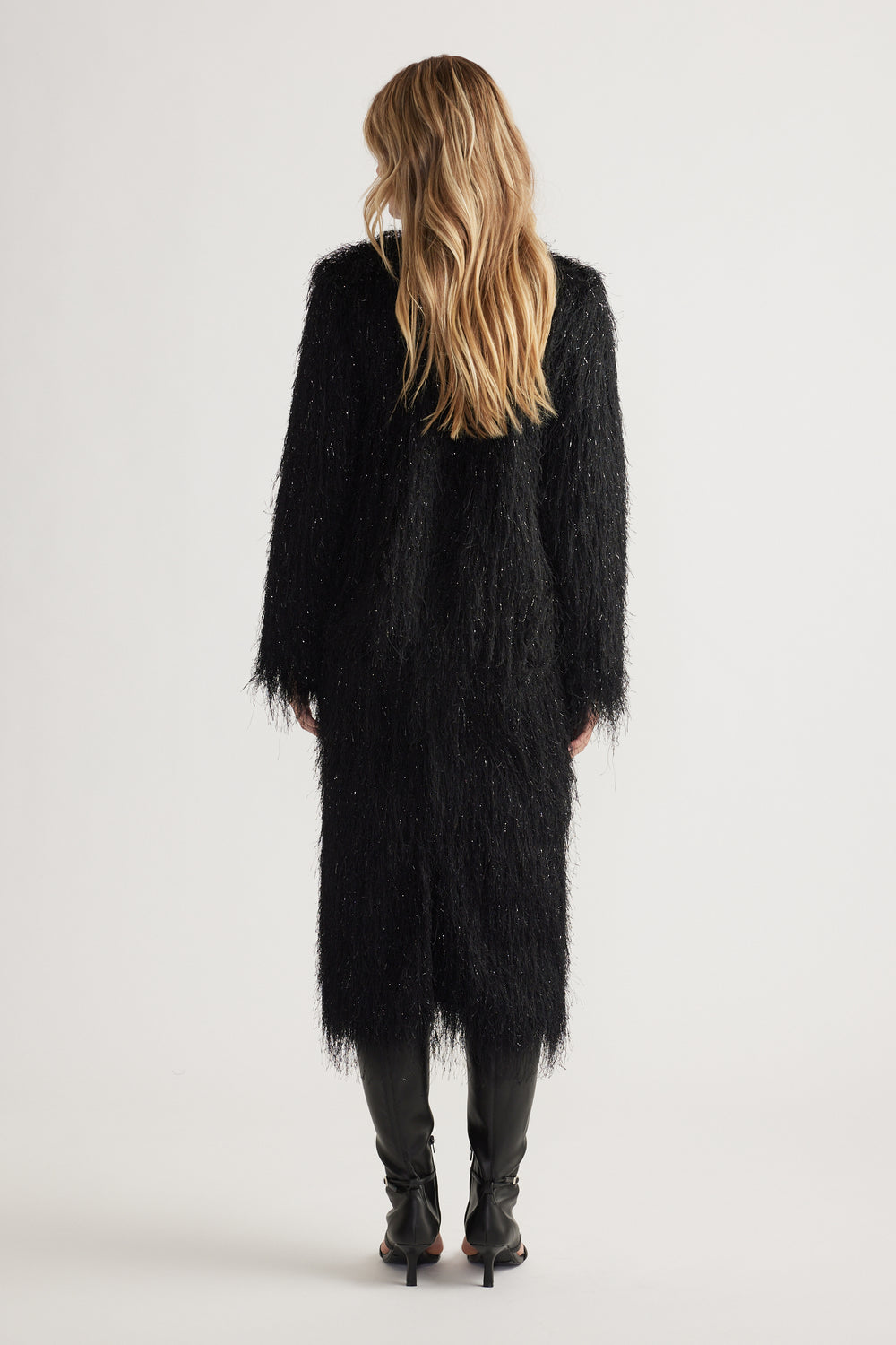 Woman seen from behind wearing a long black fringed jacket with subtle shimmer paired with black heeled boots.