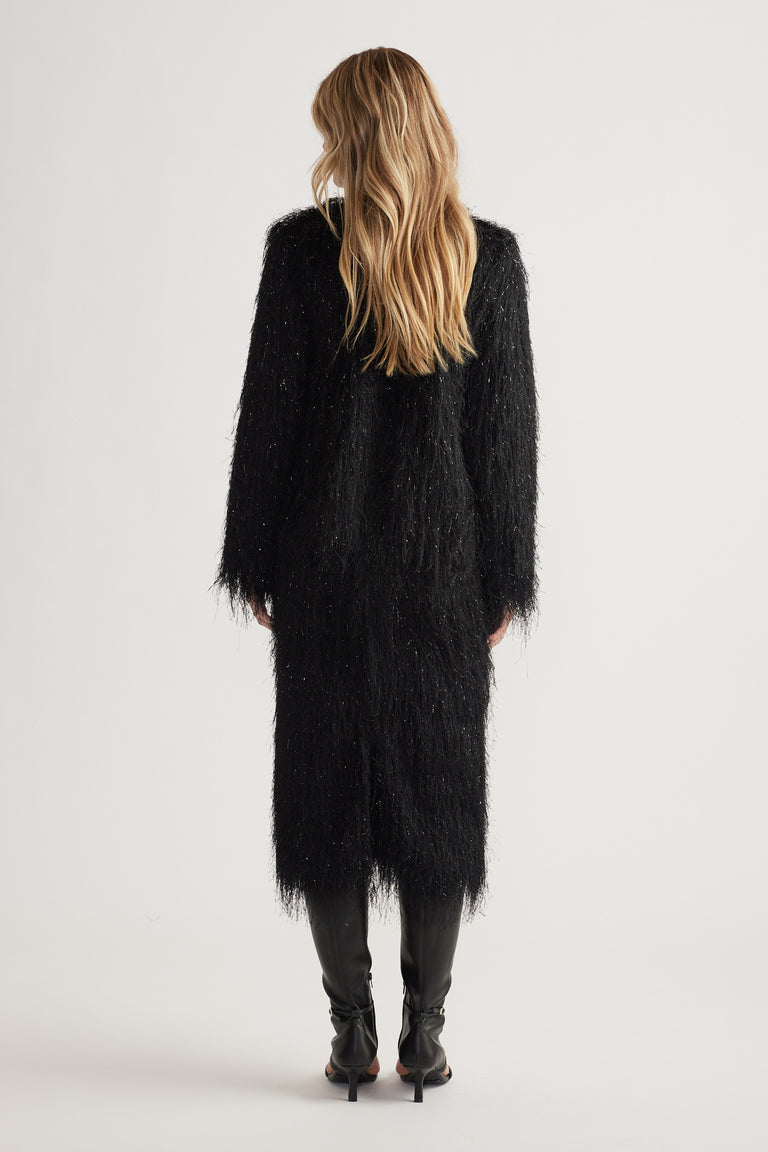 Woman seen from behind wearing a long black fringed jacket with subtle shimmer paired with black heeled boots.