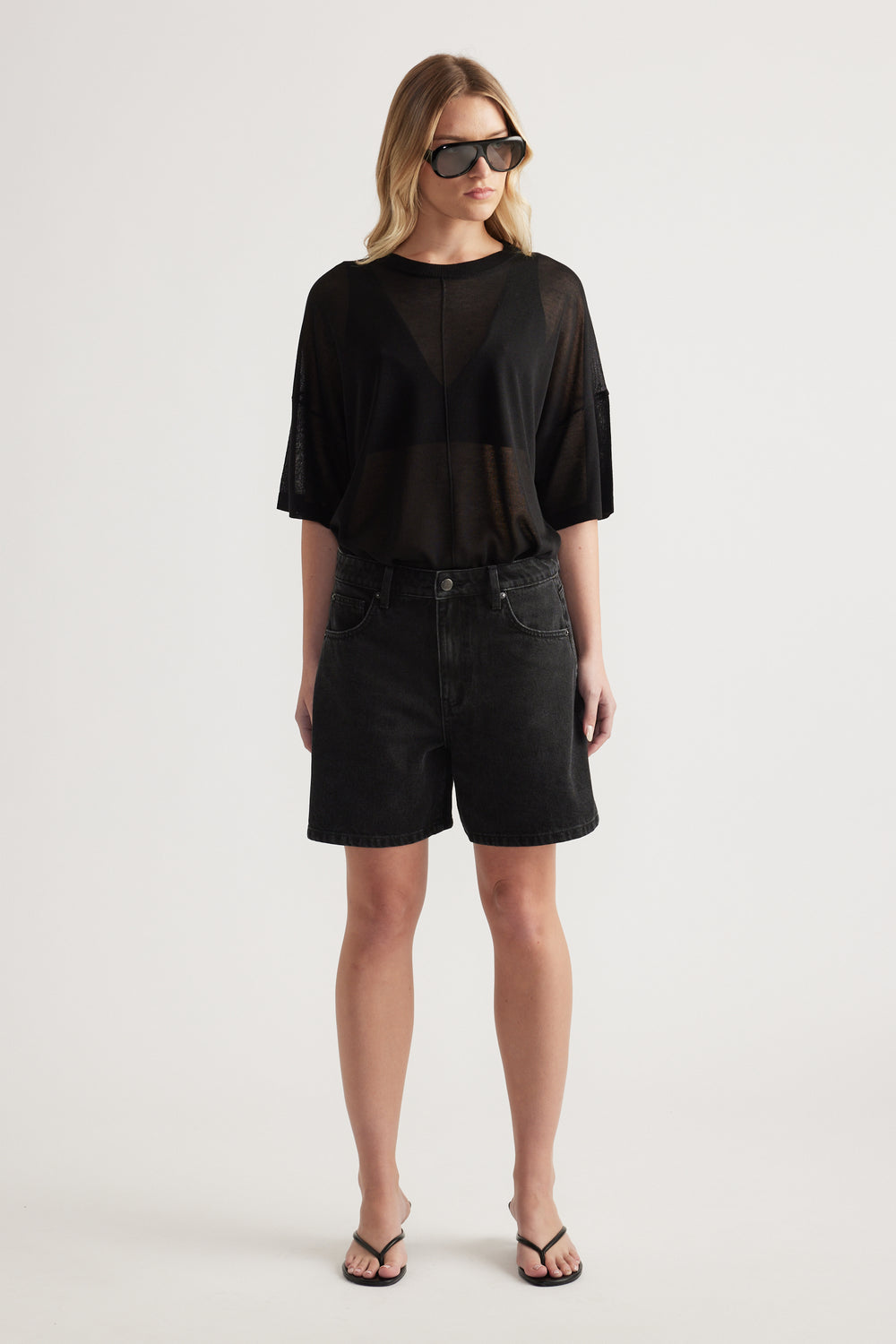 Woman wearing black high-rise A-line denim shorts with a sheer black tee and flat sandals