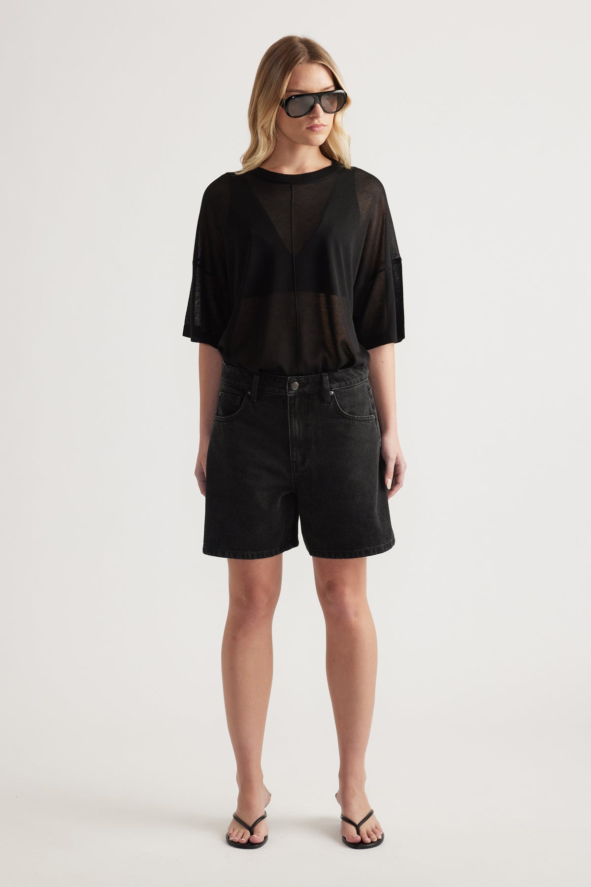 Woman wearing black high-rise A-line denim shorts with a sheer black tee and flat sandals