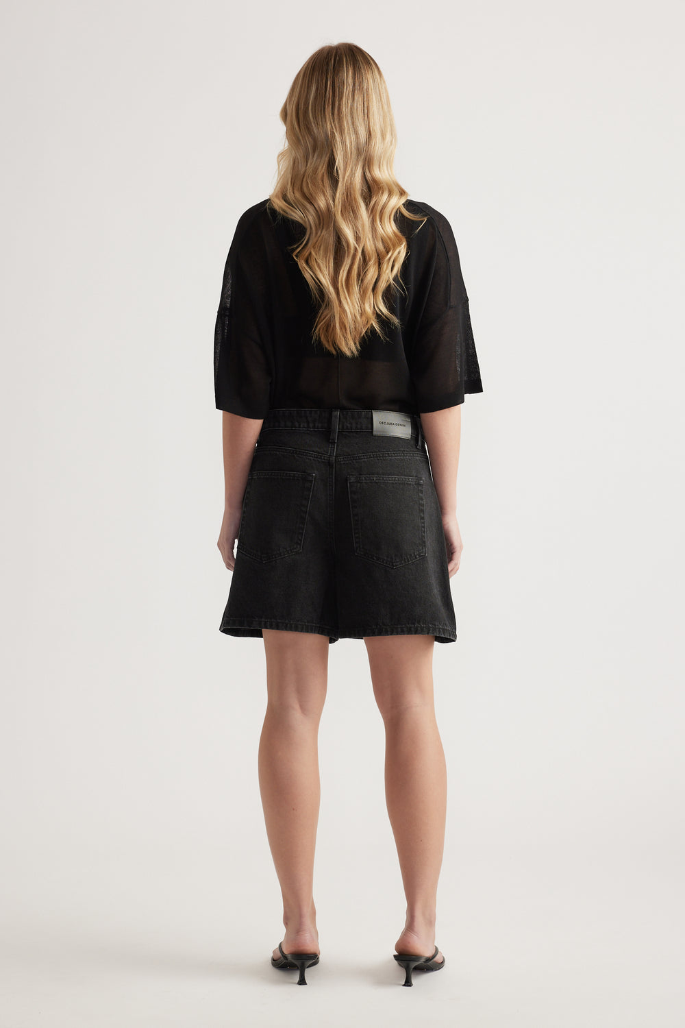 Back view of a woman in high-rise black A-line denim shorts, an oversized black tee and heeled sandals